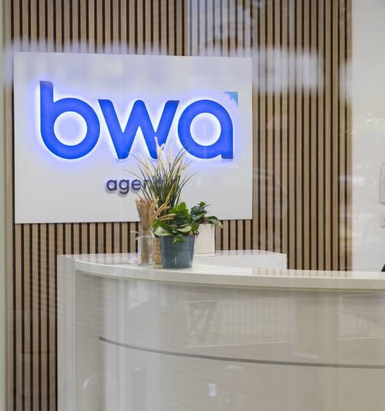 Reception avec un panneau lumineux affichant le logo 'bwa' et un comptoir blanc décoré de plantes vertes, dans un espace moderne.