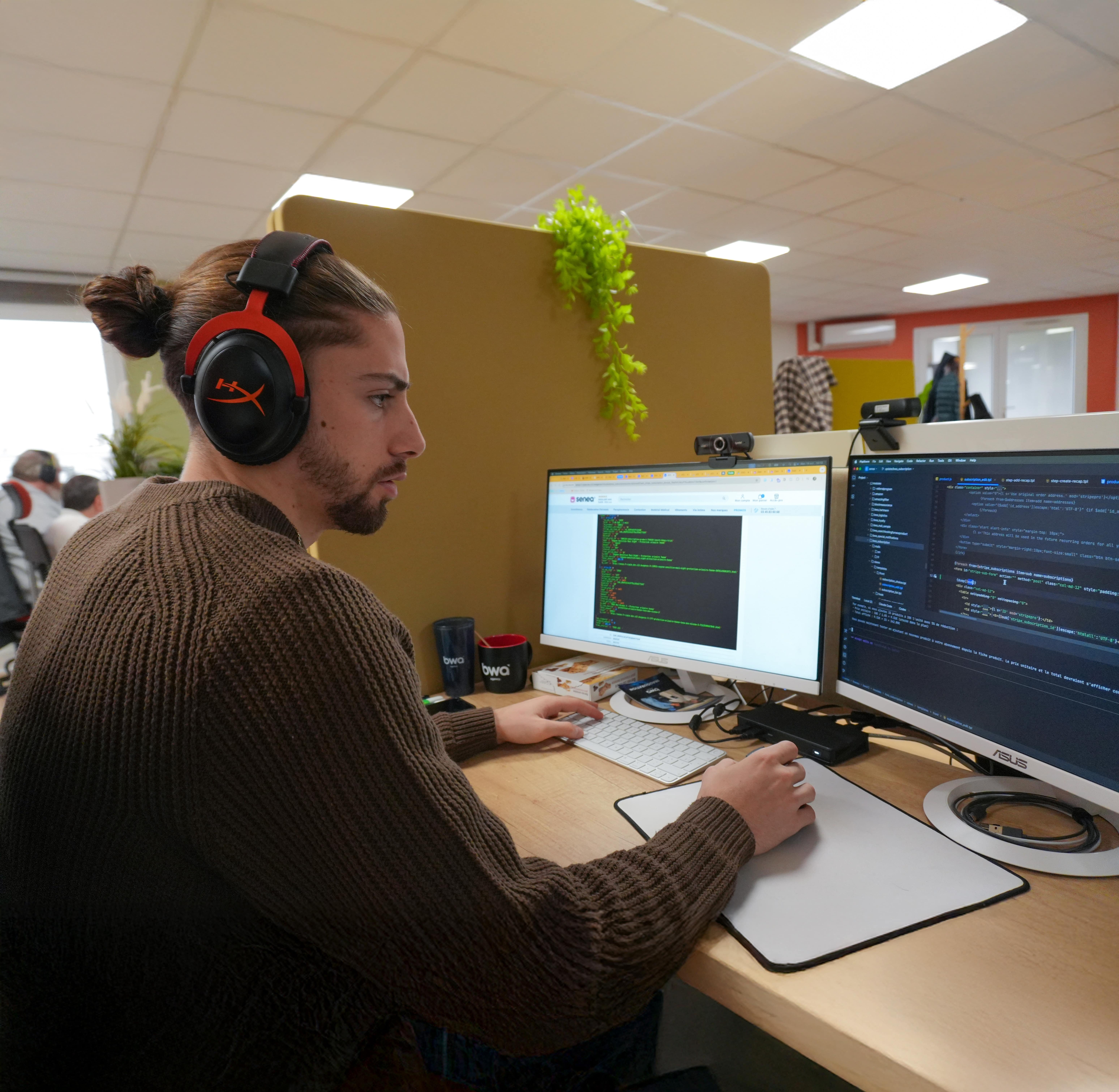 Jeune homme concentré travaillant sur deux écrans avec code informatique dans un espace de coworking, portant un casque audio rouge et marron.